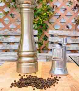 Two peppermills on a wooden table outside with vines in the background and tellicherry peppercorns on the table.