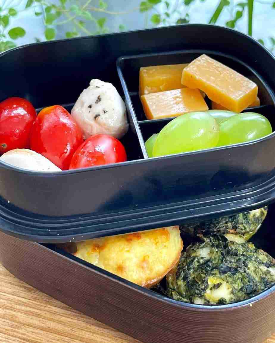 A bento lunch box filled with cherry tomatoes, mozzarella balls, grapes and gouda and mini bouchées